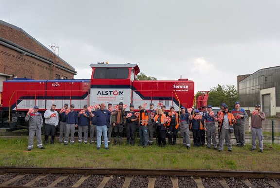 Gruppenfoto im Freien: Etwa 25 Personen stehen nebeneinander auf Gras vor einem roten Schienenfahrzeug mit blau-weißen Streifen. Auf dem Fahrzeug sind die gut lesbaren Schriftzüge „ALSTOM“, „Lokomotiven Service“, „Zürich“ und „Service ist unser Antrieb“ angebracht. Die Personen tragen überwiegend Arbeitskleidung in Grau und Blau, einige tragen orangefarbene Warnwesten und Schutzhelme. Viele halten kleine rote Schilder mit weißem Symbol oder Schrift in die Kamera. Im Vordergrund verlaufen Eisenbahnschienen. Im Hintergrund sind Backstein- und Industriegebäude sowie ein bewölkter Himmel zu sehen.
