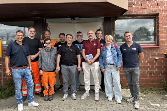 Elf Personen stehen vor dem Eingang eines Backsteingebäudes und posieren für ein Gruppenfoto. Die Personen tragen unterschiedliche Kleidung, darunter dunkle Poloshirts, T‑Shirts, eine Jeansjacke, eine graue Kapuzenjacke sowie mehrere orangefarbene Arbeitshosen mit Reflexstreifen. Im Hintergrund sind eine Doppeltür, Fenster und die Backsteinfassade des Gebäudes zu sehen. Der Boden besteht aus Pflastersteinen, der Himmel spiegelt sich in den Fensterscheiben.