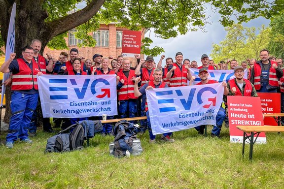 Gruppe von etwa 20–25 Personen im Freien auf einer Rasenfläche unter einem großen Baum. Viele Personen tragen rote Warnwesten und halten weiße Banner mit blau-rotem Logo und der Aufschrift „EVG Eisenbahn- und Verkehrsgewerkschaft“. Mehrere Personen zeigen erhobene Fäuste. Im Hintergrund steht ein rotes Backsteingebäude mit Fenstern. Rechts und mittig sind rote Schilder mit weißer Schrift zu sehen: „Heute ist kein Arbeitstag. Heute ist STREIKTAG!“. Vor der Gruppe liegen Taschen und Jacken auf dem Boden.