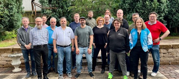Gruppenfoto mit etwa 15 Personen, die im Freien vor einer Mauer und Bäumen in zwei Reihen stehen und in die Kamera schauen. Die Personen stehen dicht nebeneinander auf einem gepflasterten Boden. In der vorderen Reihe stehen mehrere Personen mit verschiedenen Oberteilen wie Hemden, T‑Shirts und einer Jacke; rechts trägt eine Person eine auffällige blaue Weste. In der hinteren Reihe stehen weitere Personen leicht erhöht. Im Hintergrund sind grüne Sträucher und Bäume sowie eine helle Wand zu sehen. Links am Bildrand steht ein dekorativer Stein- oder Betonpflanztopf, dahinter ist eine Metallskulptur erkennbar. Die Kleidung ist überwiegend alltagstauglich, mit Jeans und schlichten Schuhen.