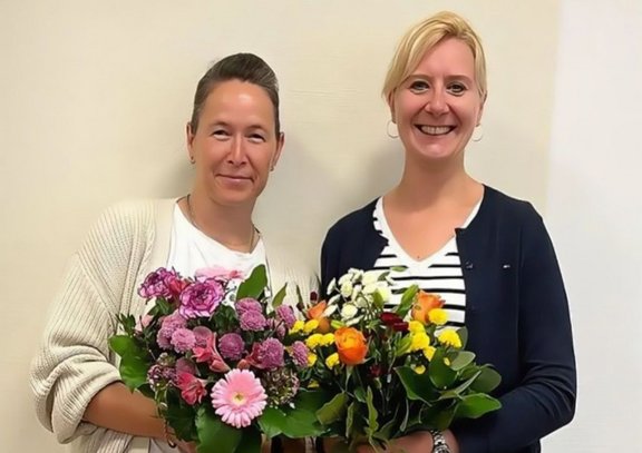 Zwei Frauen stehen nebeneinander vor einer hellen Wand und halten jeweils einen Blumenstrauß mit bunten Blüten und grünen Blättern in den Händen; beide blicken in die Kamera und lächeln.
