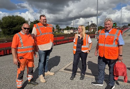 Vier Personen in orangefarbenen Warnwesten stehen auf einem erhöhten Bereich neben Bahngleisen. Im Hintergrund sind Gleisanlagen, Gebäude, ein Ampelsignal sowie ein bewölkter Himmel sichtbar.