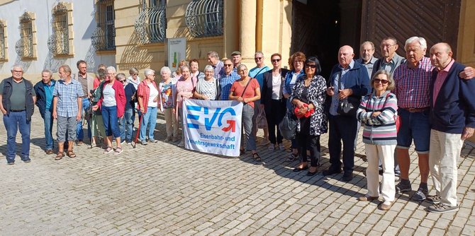 Gruppe von etwa 30 Personen steht in einer Reihe auf einem gepflasterten Platz vor einem historischen Gebäude mit gelber Fassade, hohen Fenstern und schmiedeeisernen Fenstergittern. In der Mitte halten mehrere Personen ein weißes Banner mit blauem Logo und dem gut lesbaren Text „EVG“ sowie darunter „Eisenbahn- und Verkehrsgewerkschaft“. Die Personen tragen unterschiedliche Alltagskleidung und Taschen; einige Gesichter sind unscharf dargestellt. Sonnenlicht wirft Schatten auf den Boden.
