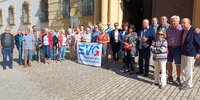Gruppe von etwa 30 Personen steht in einer Reihe auf einem gepflasterten Platz vor einem historischen Gebäude mit gelber Fassade, hohen Fenstern und schmiedeeisernen Fenstergittern. In der Mitte halten mehrere Personen ein weißes Banner mit blauem Logo und dem gut lesbaren Text „EVG“ sowie darunter „Eisenbahn- und Verkehrsgewerkschaft“. Die Personen tragen unterschiedliche Alltagskleidung und Taschen; einige Gesichter sind unscharf dargestellt. Sonnenlicht wirft Schatten auf den Boden.