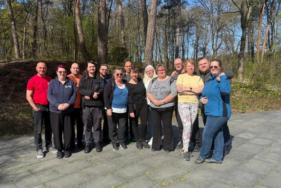 Gruppe von etwa 15 Personen steht nebeneinander auf einem gepflasterten Weg im Freien. Im Hintergrund sind hohe Bäume und Wald zu sehen. Die Personen tragen unterschiedliche Freizeitkleidung. Tageslicht, klarer Himmel.