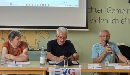 Drei ältere Personen sitzen an einem Tisch vor einer Wand mit projizierter Präsentation. Eine Person rechts hält ein Mikrofon und spricht, die beiden anderen schauen auf Unterlagen vor sich. Auf dem Tisch stehen Gläser, eine Flasche Wasser und Papiere, vorne hängt ein Banner mit der Aufschrift „EVG“.