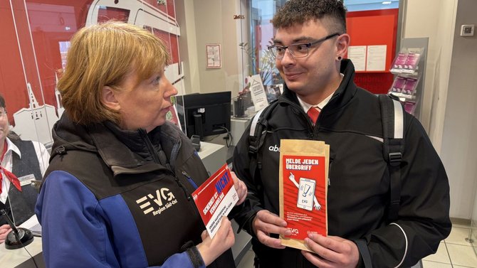 Zwei Personen stehen in einem Innenraum mit roter und weißer Gestaltung. Beide tragen dunkle Jacken mit EVG‑Aufdruck. Die Person links hält einen roten und weißen Flyer, die Person rechts hält einen roten Flyer mit der Aufschrift „Melde jeden Übergriff“. Im Hintergrund sind ein Schalterbereich, Informationsaushänge und weitere Personen zu sehen.
