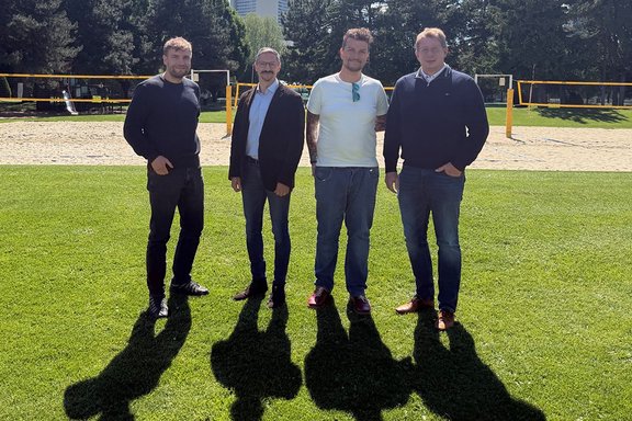 Vier Personen der EVG-Delegation stehen im Freien auf Rasen und schauen in Richtung Kamera. Hinter ihnen befindet sich ein Sandfeld, das durch ein gelbes Volleyball-Netz und Pfosten begrenzt ist. Im Hintergrund stehen viele hohe Bäume, dahinter ragt ein hohes Gebäude über die Baumkronen hinaus. Die Sonne scheint stark, dadurch fallen lange Schatten der Personen auf den Rasen. Die Personen tragen Freizeitkleidung wie Jeans, Pullover, Jacke und T‑Shirt.