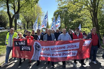 Gruppe von Menschen auf einer Straße, gesäumt von Bäumen mit grünem Laub. Im Vordergrund wird ein rotes Transparent getragen mit den gut lesbaren Texten „1. Mai 2026“ und „Erst unsere Jobs, dann eure Profite“ sowie einem DGB‑Logo. Dahinter sind zahlreiche weiße Fahnen mit blauen Aufdrucken und erkennbaren Gewerkschaftslogos zu sehen. Die Straße und der Gehweg verlaufen durch eine grüne, parkähnliche Umgebung.