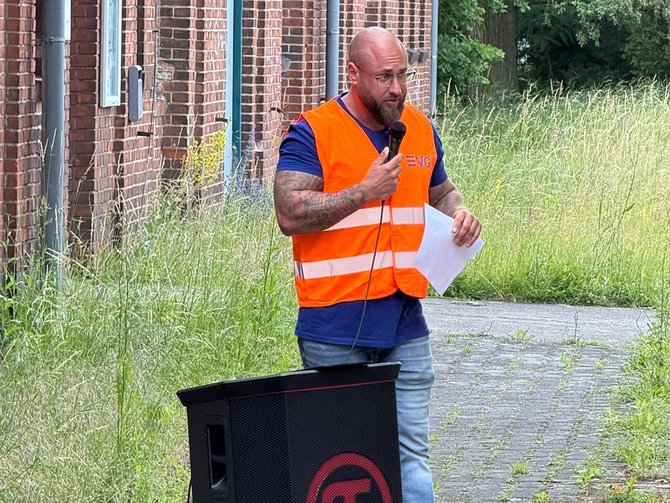 Eine Person mit orangefarbener Warnweste spricht draußen vor einem Gebäude in ein Mikrofon und hält ein Blatt Papier in der Hand. Die Szene spielt im Freien neben einem Backsteingebäude. Im Vordergrund steht eine Person mit kurz geschnittenem Haar, Brille und sichtbaren Tätowierungen an den Armen. Die Person trägt eine orangefarbene Warnweste mit der Aufschrift „EVG“ über einem dunklen T‑Shirt und spricht in ein Mikrofon, das mit der anderen Hand gehalten wird. In der Hand befindet sich außerdem ein Blatt Papier, vermutlich mit Notizen oder einer Rede. Vor der Person steht ein großer Lautsprecher auf dem Boden. Der Untergrund ist ein gepflasterter Weg, der von hohem Gras und Pflanzen gesäumt ist. Im Hintergrund sind Teile der Gebäudefassade, Fenster und Grünflächen zu sehen.