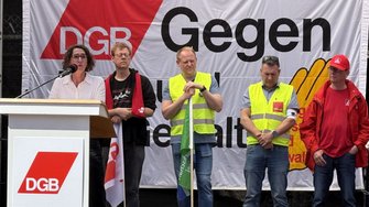 Mehrere Personen stehen hinter einem Rednerpult vor einem großen Banner. Auf dem Banner sind die Buchstaben „DGB“ sowie das Wort „Gegen“ sichtbar. Einige Personen tragen Warnwesten oder Jacken. Vor ihnen steht ein Mikrofon, der Hintergrund ist das Banner.
