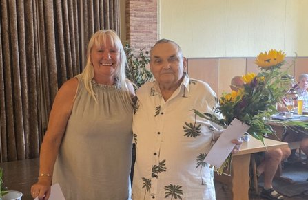 Zwei Personen stehen nebeneinander vor einem Vorhang. Eine Person hält einen Blumenstrauß mit Sonnenblumen und einen weißen Umschlag. Im Hintergrund sind weitere Personen an Tischen zu sehen.