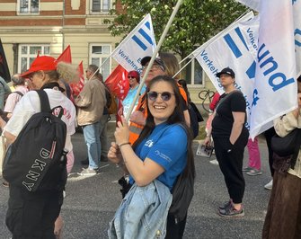 Menschen stehen auf einer Straße und halten Fahnen. Im Vordergrund hält eine Person eine weiße Fahne mit blauem Schriftzug „EVG Verkehrsgewerkschaft“. Weitere weiße und rote Fahnen sind sichtbar. Mehrere Personen tragen Rucksäcke oder Warnwesten. Im Hintergrund stehen ein mehrstöckiges Gebäude mit Fenstern, Bäume mit grünem Laub und weitere Teilnehmende.