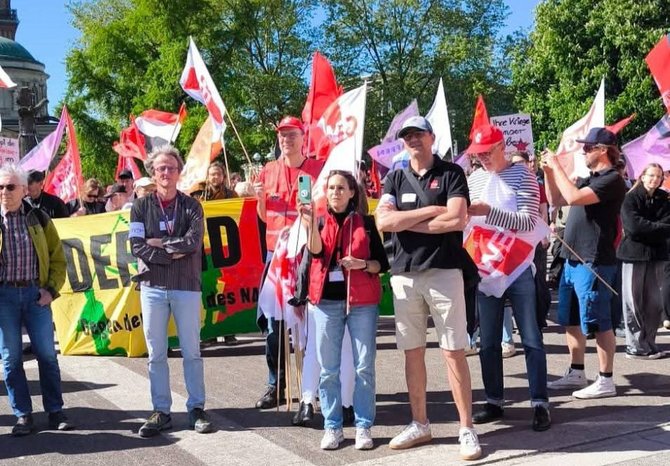 Eine Gruppe von Menschen steht auf einer Straße, viele tragen rote Fahnen oder halten Fahnenstangen. Im Vordergrund stehen mehrere Personen nebeneinander, teils mit verschränkten Armen. Im Hintergrund sind weitere Menschen, Bäume und Fahnen zu sehen. Ein großes gelb-grünes Banner mit schwarzer Schrift ist teilweise sichtbar, der genaue Text ist nicht vollständig erkennbar.