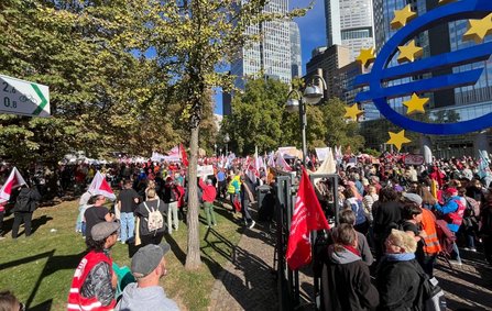 Große Versammlung im Freien vor Hochhäusern. Rechts steht ein großes blaues Euro-Symbol mit gelben Sternen. Zahlreiche Menschen befinden sich auf einer Grünfläche und einem Weg, viele tragen oder halten rote und weiße Fahnen und Banner. Bäume und moderne Bürogebäude bilden den Hintergrund.