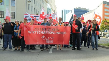 Gruppe von Demonstrationsteilnehmenden auf einer städtischen Straße. Vorne wird ein großes rotes Banner mit der Aufschrift „ZEIT HEILT KEINE WIRTSCHAFTSWUNDEN.“ getragen. Dahinter sind Fahnen, Schilder und weitere Teilnehmende zu sehen. Im Hintergrund stehen mehrstöckige Gebäude und Bäume.