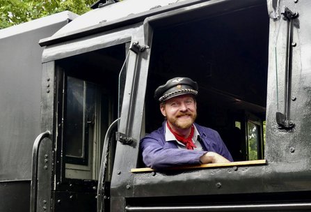 Schwarzer Lokomotivführerstand mit geöffnetem Seitenfenster. Eine Person in dunkler Uniformmütze und Jacke lehnt sich auf die Fensterkante. Sichtbar sind die genieteten Metallflächen der Lokomotive sowie ein geöffneter Zugang hinter der Person.