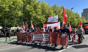 Demonstrationszug auf einer Straße bei sonnigem Wetter. Mehrere Personen halten ein langes rotes Banner mit der Aufschrift „Erst unsere Jobs, dann eure Profite“, daneben „1. Mai 2026“ und „DGB“. Um das Banner herum werden rote Fahnen getragen. Im Hintergrund stehen Bäume und mehrstöckige Gebäude.