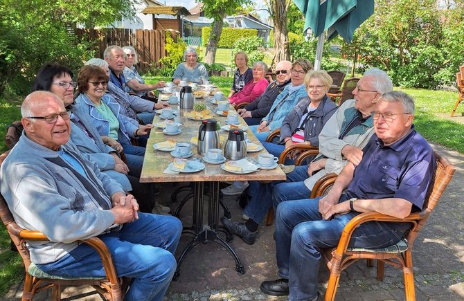 Eine große Gruppe von etwa zwölf bis fünfzehn Personen sitzt an einem langen rechteckigen Tisch im Freien in einem Garten. Der Tisch ist mit mehreren Tassen, Untertassen, Kaffeekannen und Kuchentellern gedeckt. Die Personen sitzen auf beiden Seiten des Tisches auf Stühlen und schauen in Richtung Kamera. Im Hintergrund sind Bäume, Sträucher, ein Holzzaun und ein Gebäude zu sehen. Das Licht ist hell, und der Bereich ist teilweise von Schatten durch Bäume bedeckt.
