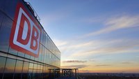 Großes rotes „DB“-Logo an der verglasten Fassade eines Hochhauses; im Hintergrund eine weite Stadtansicht unter einem Himmel mit Abendfarben und vereinzelten Wolken.