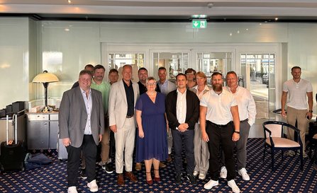 Gruppenfoto in einem großen Raum mit Teppichboden, Stühlen und einem Sideboard. Mehrere Personen stehen in zwei Reihen vor einer Glaswand mit Türen. Im Hintergrund sind weitere Personen, Möbelstücke und ein beleuchtetes Notausgangsschild zu sehen.