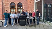 Elf Personen stehen auf einem gepflasterten Innenhof hinter einem schwarzen Gasgrill und einem Servierwagen mit Tellern. Im Hintergrund befindet sich ein Gebäude aus rotem Backstein mit mehreren hohen Rundbogenfenstern. Rechts ist eine Glasfassade mit Spiegelungen zu sehen, daneben stehen Fahrräder an einem Fahrradständer. Die Personen tragen überwiegend Freizeitkleidung und sind in einer Reihe um den Grill gruppiert. Tageslicht, keine sichtbaren Texte im Bild.
