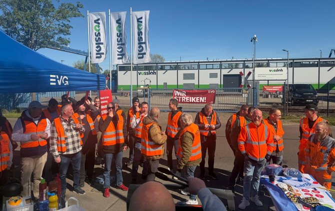 Eine Gruppe von Menschen trägt orange Warnwesten und steht vor einem Industriegelände mit Gebäuden und Fahnen mit der Aufschrift „Vossloh“. Mehrere Schilder sind sichtbar, darunter ein rotes Schild mit der Aufschrift „WIR STREIKEN!“. Ein blauer Pavillon mit EVG‑Logo steht links im Bild.