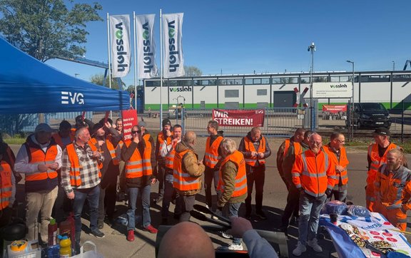 Eine Gruppe von Menschen trägt orange Warnwesten und steht vor einem Industriegelände mit Gebäuden und Fahnen mit der Aufschrift „Vossloh“. Mehrere Schilder sind sichtbar, darunter ein rotes Schild mit der Aufschrift „WIR STREIKEN!“. Ein blauer Pavillon mit EVG‑Logo steht links im Bild.