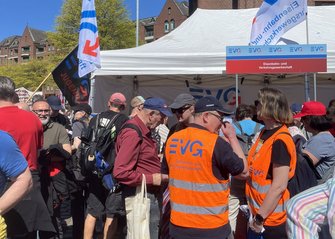 Menschenmenge vor einem weißen Pavillon. Am Pavillon ist ein Schild mit der Aufschrift „EVG Eisenbahn- und Verkehrsgewerkschaft“ angebracht. Mehrere Personen tragen orange Warnwesten mit blauem „EVG“-Schriftzug. Weiße Fahnen mit blauem „EVG“-Logo und eine schwarze Fahne mit rotem und blauem Aufdruck sind zu sehen. Im Hintergrund stehen Backsteingebäude, Bäume und weitere Fahnen.