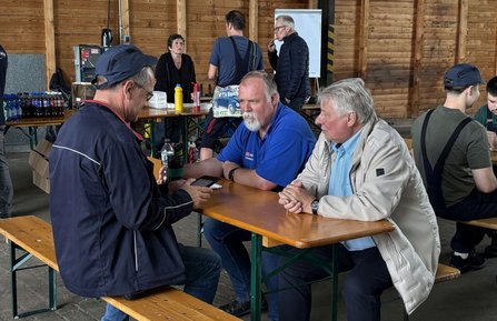 Mehrere Personen sitzen und stehen an Holzbänken und Tischen in einer großen Halle mit Holzwänden. Drei Personen sitzen an einem Tisch im Vordergrund und schauen auf ein Mobiltelefon. Auf einem Tisch im Hintergrund stehen Getränkeflaschen und Lebensmittel. Weitere Personen befinden sich im Gespräch oder sitzen an Bänken.