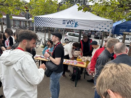 Mehrere Personen stehen an einem Informationsstand unter einem weißen Pavillon mit der Aufschrift „EVG JUGEND“. Auf Holztischen liegen Stofftaschen, rote Flyer, Schlüsselbänder, kleine verpackte Gegenstände und weitere Informationsmaterialien. Einige Personen greifen nach Materialien oder unterhalten sich. Im Hintergrund sind Fahrräder, parkende Autos, Bäume und Gebäude zu sehen.