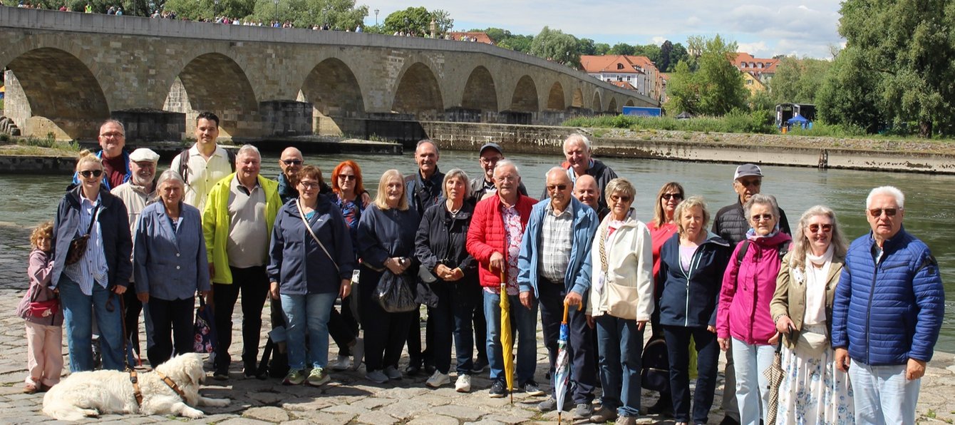Eine größere Gruppe von Menschen steht gemeinsam am Ufer eines Flusses vor einer langen Steinbrücke. Im Vordergrund stehen etwa zwanzig Personen dicht beieinander auf einem gepflasterten Weg am Wasser. Einige halten Spazierstöcke oder tragen Rucksäcke und Jacken. Ein Hund liegt vorne am Boden. Hinter der Gruppe fließt ein breiter Fluss. Darüber verläuft eine lange, historische Steinbrücke mit vielen Bögen, auf der ebenfalls mehrere Menschen stehen.