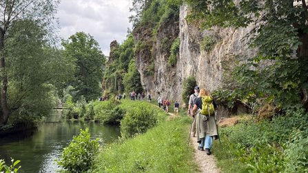 Mehrere Personen wandern auf einem schmalen Pfad am Rand eines ruhigen Gewässers entlang. Rechts erhebt sich eine steile, felsige Wand mit Bewuchs, links fließt das Wasser. Einige tragen Rucksäcke und gehen hintereinander.