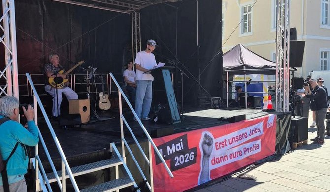 Eine Bühne im Freien mit einem Rednerpult. Eine Person steht am Pult und hält ein Blatt Papier. Links auf der Bühne sitzen weitere Personen mit Musikinstrumenten, darunter eine Gitarre. An der Vorderseite der Bühne hängt ein rotes Banner mit der Aufschrift „1. Mai 2026“ und „Erst unsere Jobs, dann eure Profite“ sowie einer grafischen Faust. Vor der Bühne stehen Menschen, einige mit Kameras.