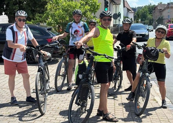Fünf Personen stehen mit Fahrrädern auf einem gepflasterten Bereich an einer Straße. Alle tragen Fahrradhelme, einige zusätzlich Sonnenbrillen. Eine Person vorne trägt ein leuchtend gelbes ärmelloses Oberteil, die anderen tragen überwiegend kurzärmlige Kleidung in unterschiedlichen Farben. Die Fahrräder stehen nebeneinander, mehrere Personen halten die Lenker fest. Im Hintergrund befinden sich geparkte Autos, ein Haus mit Zaun, eine Ampel mit grünem Signal und weitere Gebäude. Es ist sonnig, mit deutlichen Schatten auf dem Boden.