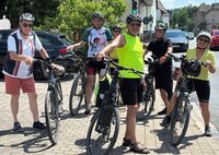 Fünf Personen stehen mit Fahrrädern auf einem gepflasterten Bereich an einer Straße. Alle tragen Fahrradhelme, einige zusätzlich Sonnenbrillen. Eine Person vorne trägt ein leuchtend gelbes ärmelloses Oberteil, die anderen tragen überwiegend kurzärmlige Kleidung in unterschiedlichen Farben. Die Fahrräder stehen nebeneinander, mehrere Personen halten die Lenker fest. Im Hintergrund befinden sich geparkte Autos, ein Haus mit Zaun, eine Ampel mit grünem Signal und weitere Gebäude. Es ist sonnig, mit deutlichen Schatten auf dem Boden.
