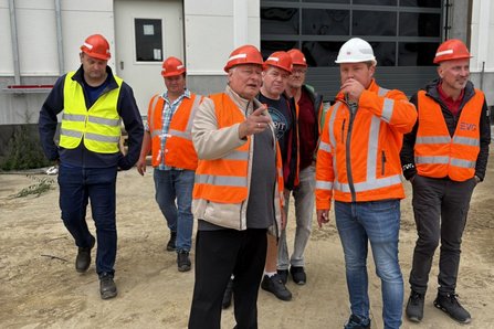 Mehrere Personen mit Schutzhelmen und Warnwesten stehen vor einem Industriegebäude mit großem Tor. Eine Person zeigt mit der Hand in die Ferne, während eine andere Person mit weißem Helm daneben steht. Der Boden besteht aus unbefestigtem Untergrund, im Hintergrund sind Gebäude- und Baustellenelemente sichtbar.