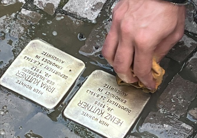 Zwei quadratische Messingtafeln sind in ein Pflaster eingelassen und teilweise von Wasser umgeben. Eine Hand reinigt eine der Tafeln mit einem Tuch. Auf den Tafeln sind die Namen „Heinz Kuttner“ und „Irma Kuttner“ sowie weitere eingravierte Textzeilen sichtbar.