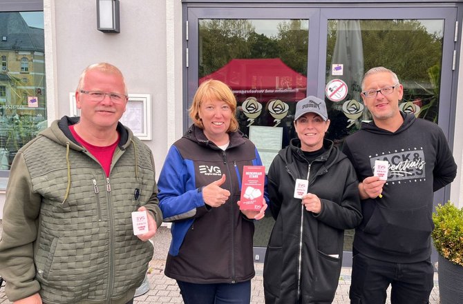 Vier Personen stehen vor einem Gebäudeeingang mit Glastür. Jede Person hält einen kleinen roten oder weißen Gegenstand mit dem Logo „EVG“, eine Person zusätzlich ein rotes Heft mit der Aufschrift „Betriebsratswahl“. Hinter der Gruppe ist die reflektierende Glasfront des Gebäudes zu sehen.