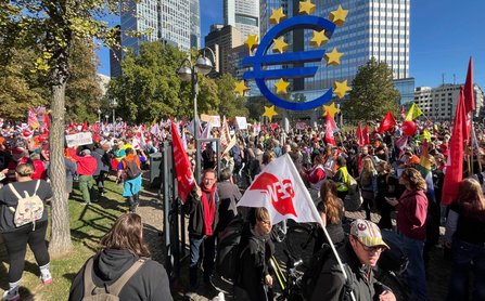 Große Menschenmenge auf einer Grünfläche und einem gepflasterten Weg vor modernen Hochhäusern. Im rechten Bildbereich steht ein großes blaues Euro-Symbol mit gelben Sternen. Viele rote und weiße Fahnen, Transparente und Schilder sind sichtbar. Bäume säumen den Platz, die Sonne scheint bei klarem Himmel.