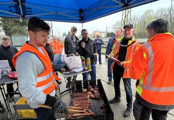 Unter einem blauen Faltpavillon stehen mehrere Personen im Freien. Einige Personen tragen orangefarbene Warnwesten, eine Person im Vordergrund trägt schwarze Grillhandschuhe und hält eine Grillzange. In der Mitte steht ein rechteckiger Grill mit mehreren Würstchen und runden Fleischstücken auf dem Rost. Daneben befindet sich ein Tisch mit Flaschen, Bechern, Behältern und weiteren Utensilien. Im Hintergrund sind weitere Personen, teilweise ebenfalls mit Warnwesten, sowie Bahngleise, Oberleitungen, Bäume und Gebäude zu sehen. Die Gesichter der Personen sind unscharf dargestellt.