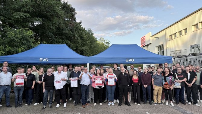 Gruppenfoto mit etwa 40 Personen auf einem gepflasterten Platz vor einem Industriegebäude mit DB-Logo. Zwei blaue Pavillons mit EVG-Aufdruck stehen hinter der Gruppe. Mehrere Personen halten rote und weiße Plakate mit Gewerkschafts- und Solidaritätsbotschaften. Im Hintergrund sind Bäume, Wolken und technische Anlagen am Gebäude zu sehen.