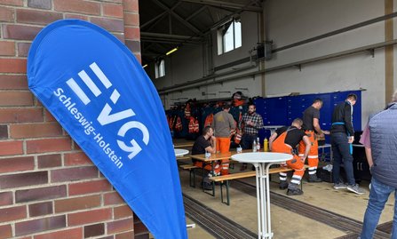 Eine blaue Fahne mit der Aufschrift „EVG Schleswig-Holstein“ steht im Vordergrund neben einer Backsteinwand. Im Hintergrund sind mehrere Personen in einer Halle zu sehen, einige tragen orange Warnkleidung. Außerdem sind Tische, Bänke, blaue Spinde und aufgehängte Warnwesten sichtbar.