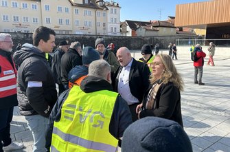 Eine Gruppe von Personen steht auf einem gepflasterten Platz im Freien. Im Vordergrund sind mehrere Personen von hinten zu sehen, darunter eine Person mit einer gelben Warnweste mit dem EVG‑Logo. Weitere Personen tragen dunkle Jacken oder Warnwesten. Im Hintergrund stehen Gebäude mit hellen Fassaden sowie ein moderner Holzbau. Einige Personen sprechen miteinander, andere gehen über den Platz. Der Himmel ist klar und sonnig.