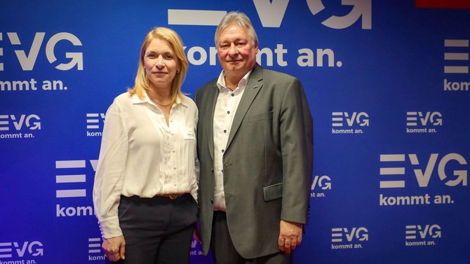 Zwei Personen stehen vor einer blauen Wand mit mehrfach sichtbarem EVG‑Logo und dem Schriftzug „kommt an.“. Die linke Person trägt ein helles Oberteil und dunkle Hose. Die rechte Person trägt einen Anzug mit hellem Hemd.