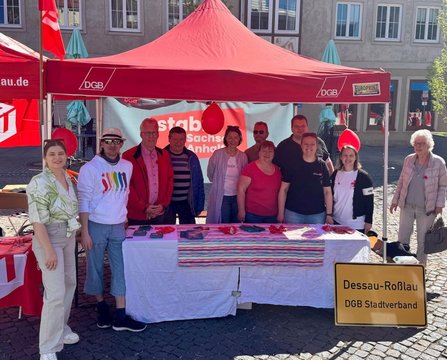 Unter einem roten Pavillon mit DGB‑Logo steht eine Gruppe von Personen hinter einem Tisch. Auf dem Tisch liegen rote Armbänder, Flyer und weitere Materialien. Am Pavillon hängen rote Luftballons. Im Hintergrund sind Gebäude mit Fenstern zu sehen. Vor dem Pavillon steht ein gelbes Schild mit der Aufschrift „Dessau‑Roßlau DGB Stadtverband“.