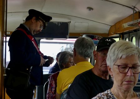 Innenraum eines historischen Zugwagens mit Holzverkleidung. Mehrere Personen sitzen auf den Sitzbänken. Im linken Vordergrund steht eine Person in Uniform und hält einen Gegenstand in den Händen. Über den Sitzen befinden sich Gepäckablagen, durch die Fenster fällt Tageslicht ein.