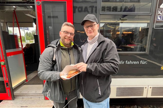Zwei Personen stehen vor einem roten Zug mit geöffneten Türen. Eine Person hält eine geöffnete Pappschale mit einem Brötchen und einer Bratwurst. Am Zug ist der Text „Wir fahren für:“ teilweise sichtbar. Im Inneren des Zuges sind Sitzplätze und weitere Personen zu erkennen.