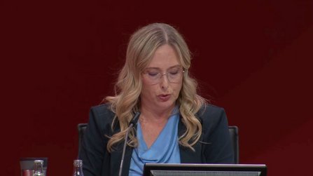 Eine Person sitzt an einem Tisch vor rotem Hintergrund. Vor der Person stehen ein Mikrofon, ein Bildschirm und mehrere Getränkeflaschen. Die Person trägt ein dunkles Jackett über einem hellen Oberteil. Der Tisch und die Technik sind frontal ausgerichtet, der Hintergrund ist einheitlich rot ohne weitere sichtbare Elemente.