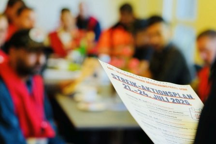 Unscharfer Hintergrund mit mehreren an Tischen sitzenden Personen in roten Warnwesten. Im Vordergrund hält eine Person ein Blatt Papier, auf dem „STREIK-AKTIONSPLAN 21.–24. JULI 2026“ lesbar ist.